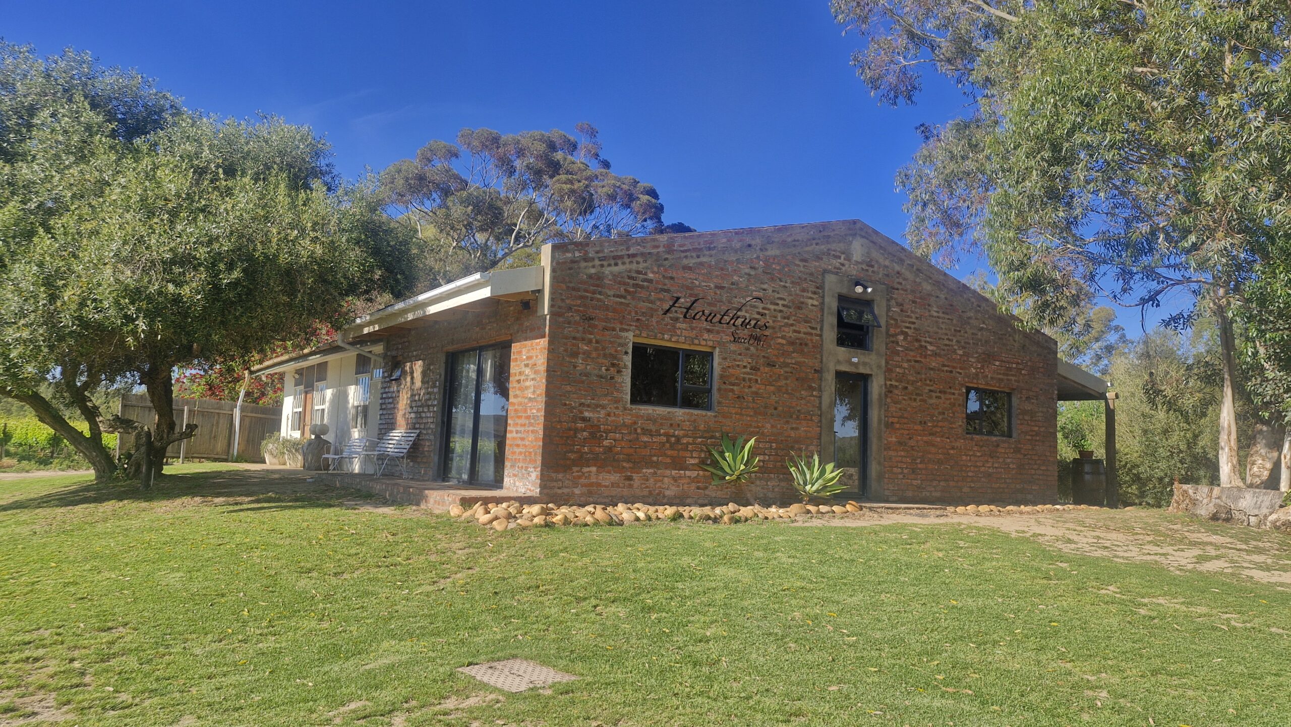 Group of people playing volleyball on a grassy area with a wooden bench labeled "DIE HOUTHUIS" in the foreground and dogs resting nearby.