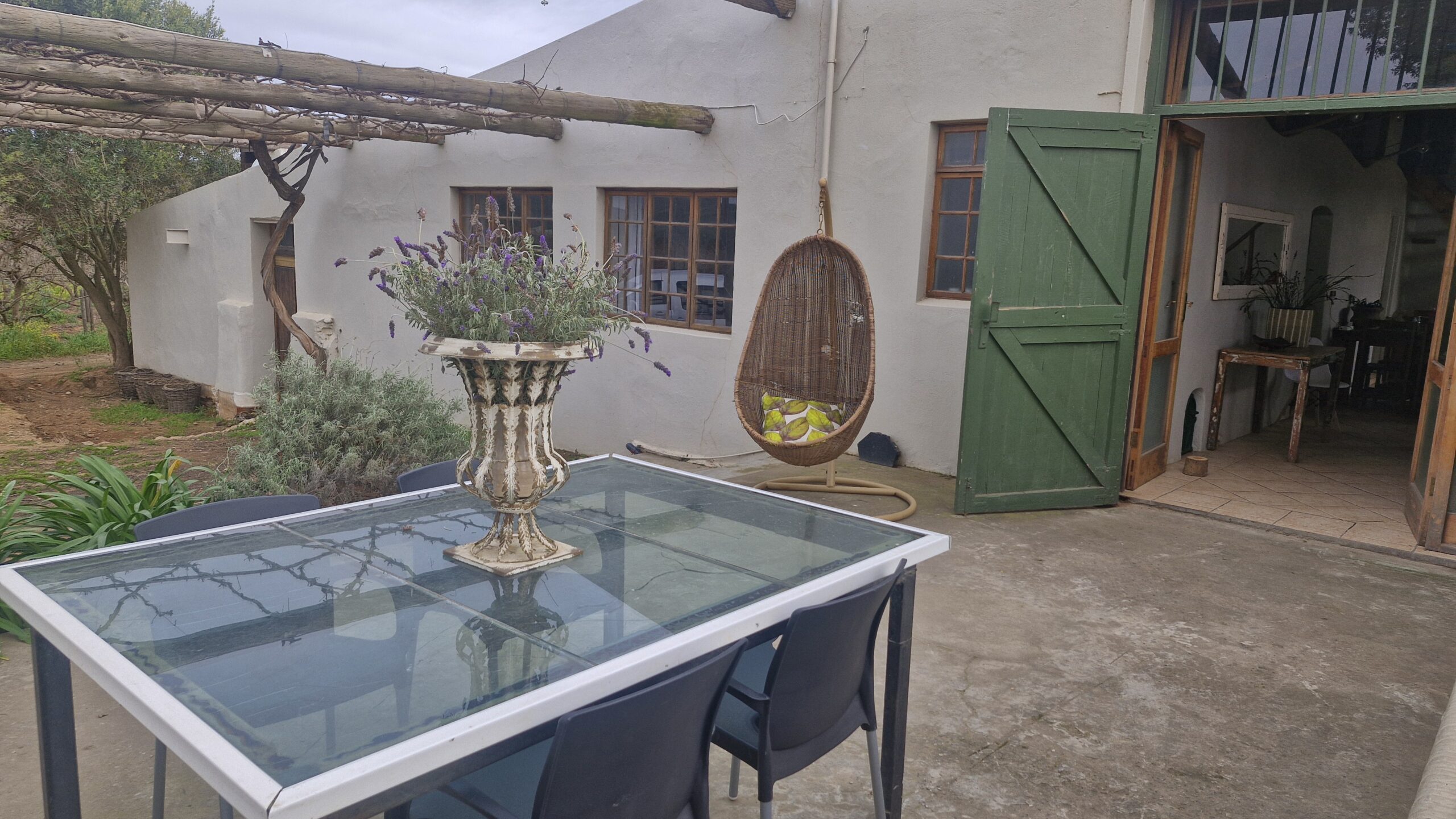 Outdoor seating area with a glass table, potted lavender, hanging wicker chair, green wooden door, and rustic building in the background.