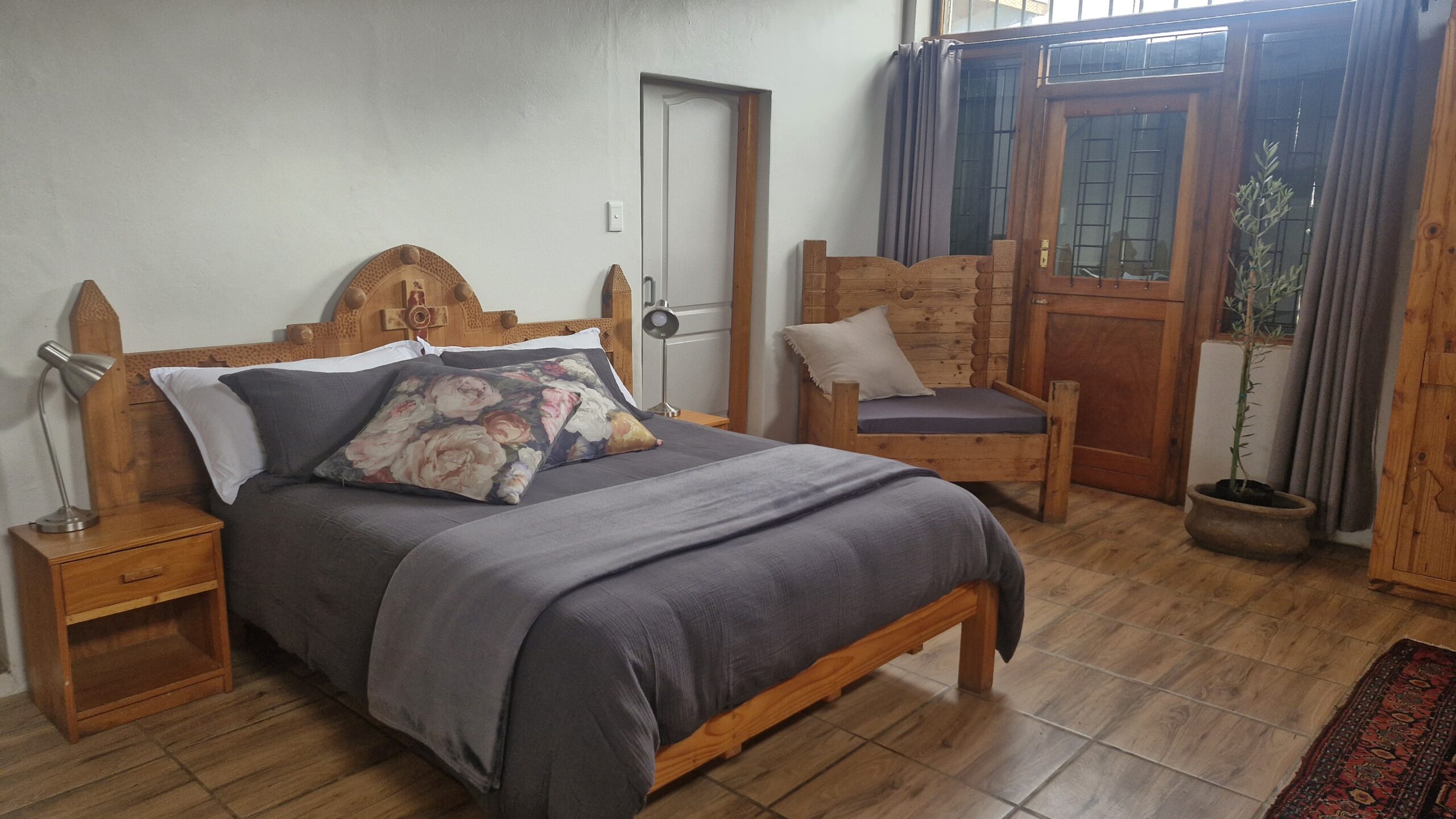Cozy bedroom with wooden furniture, floral pillows, gray bedding, and a small indoor plant near the door.